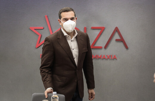 Ο Αλέξης Τσίπρας με μάσκα μπροστά από σήμα του ΣΥΡΙΖΑ